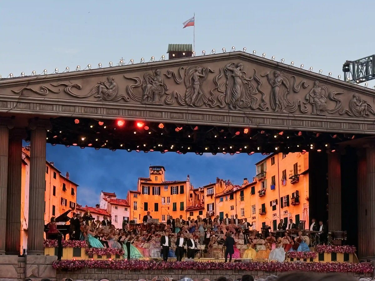 Festlich geschmückte Open-Air-Bühne mit grossem Orchester und Chor vor einer mediterranen Kulisse.