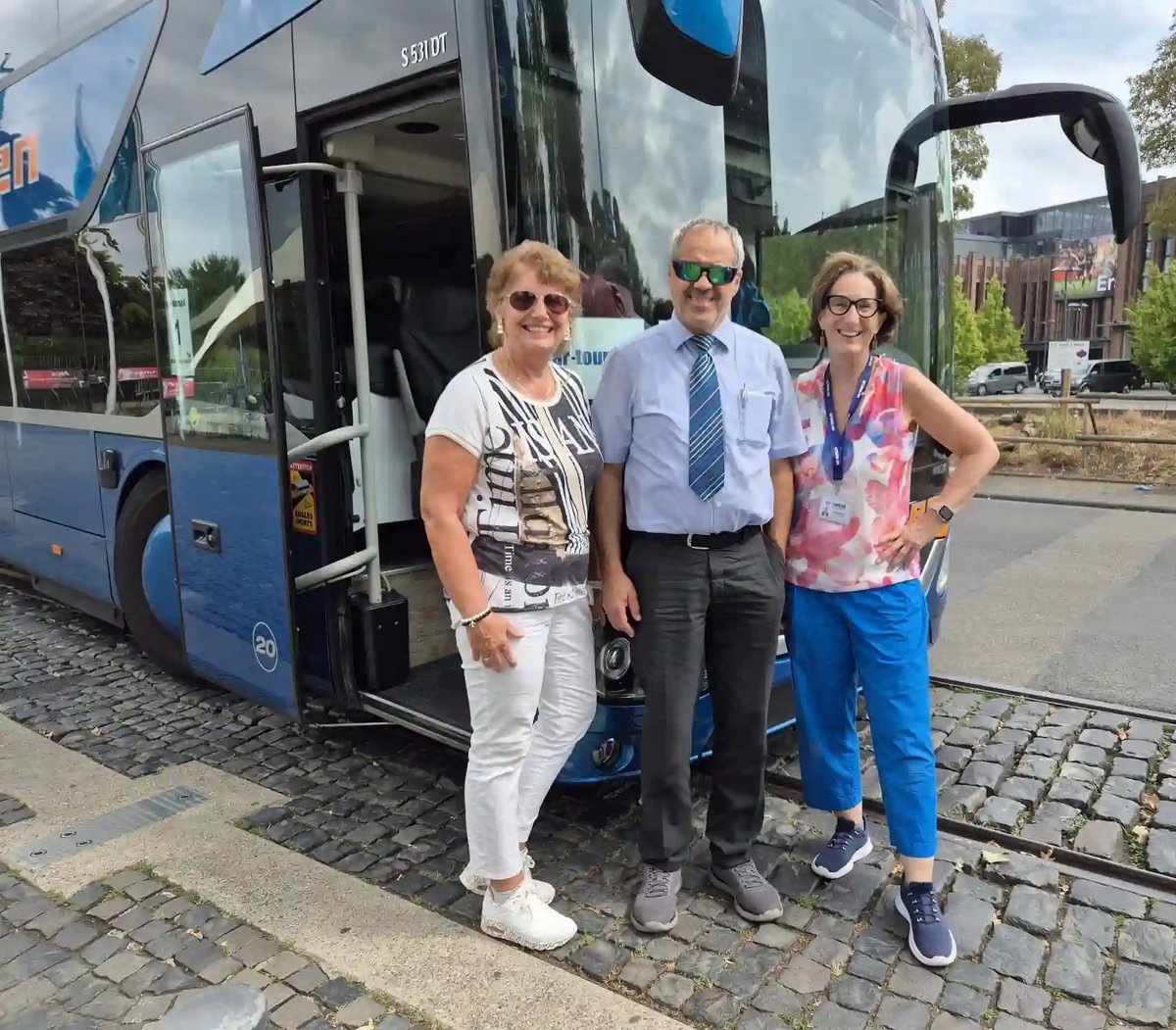 Zwei Reiseteilnehmerinnen und ein Chauffeur stehen lächelnd vor einem Reisebus.