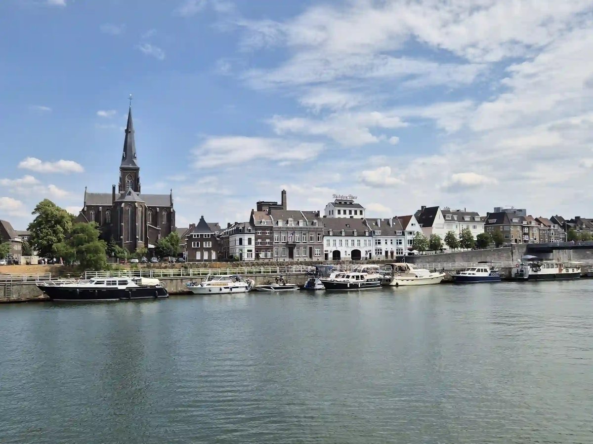 Blick über die Maas auf die Uferpromenade von Maastricht mit Booten, historischen Häusern und Kirche.