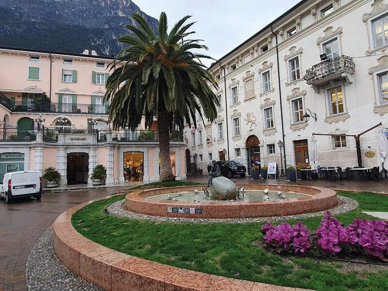 Place avec palmier, fontaine et massif de fleurs roses devant des façades historiques
