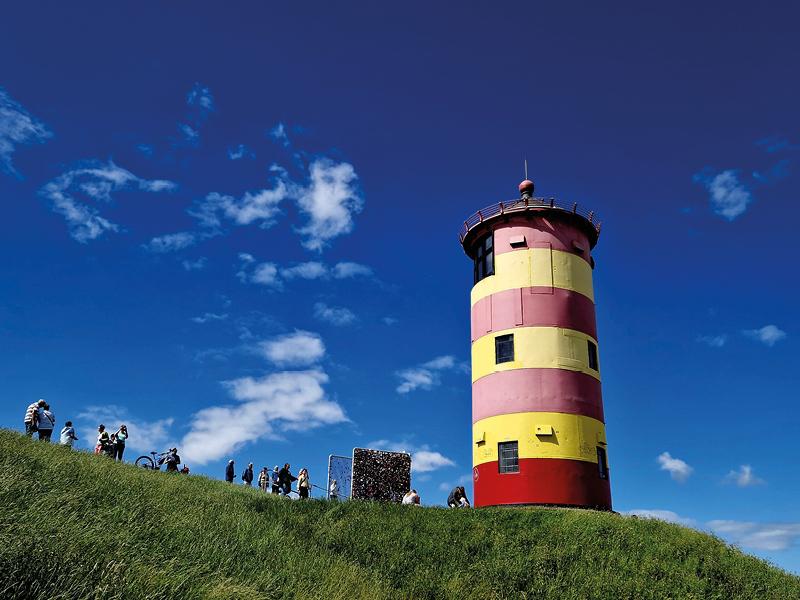 Bunter Leuchtturm auf einem Hügel mit Menschen unter blauem Himmel