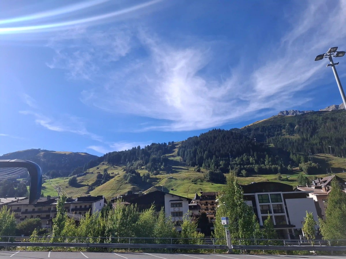 Village alpin au pied de montagnes verdoyantes, sous un ciel bleu traversé de nuages blancs