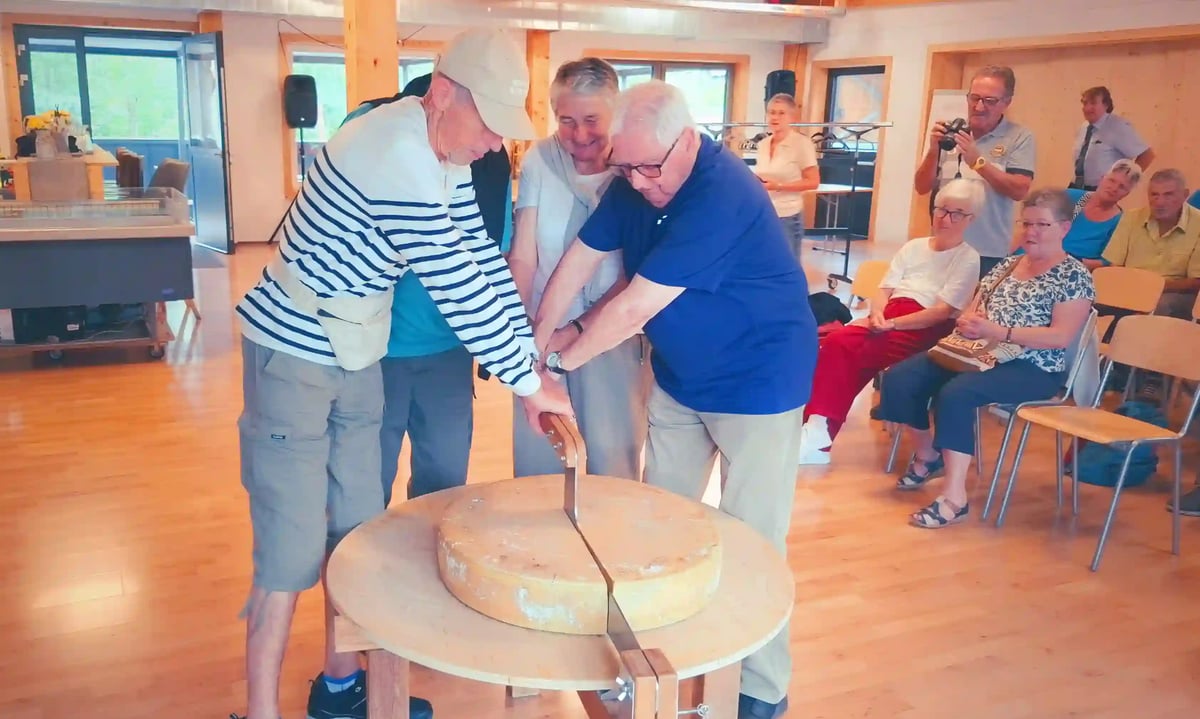 Trois voyageurs découpent une grande meule de fromage sous le regard des autres participants