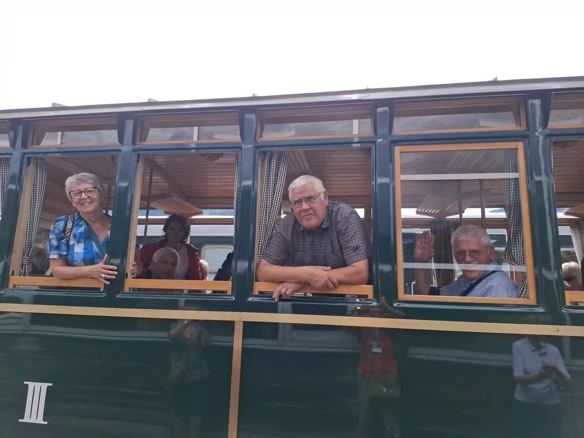 Voyageurs souriants aux fenêtres d'un wagon historique vert, dont un homme saluant de la main