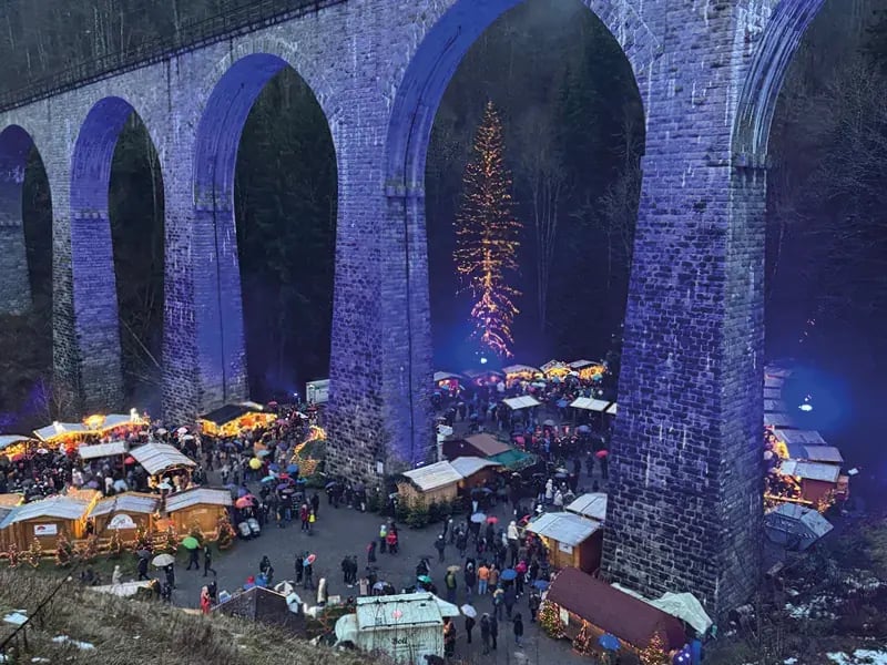 Ein stimmungsvoller Weihnachtsmarkt mit beleuchteten Holzbuden direkt unter den hohen, blau angestrahlten Steinbögen des Ravennaviadukts im Schwarzwald.