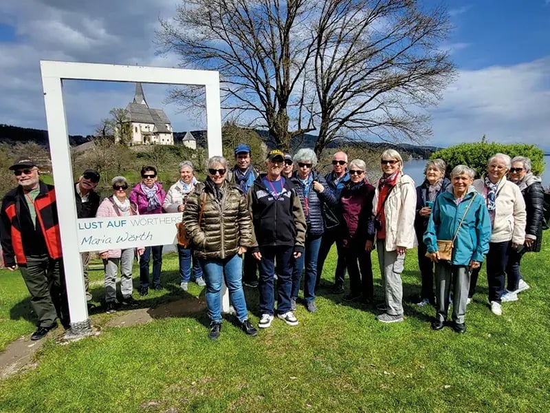 Reisegruppe posiert im Freien neben einem großen Rahmen, im Hintergrund Kirche und Bäume
