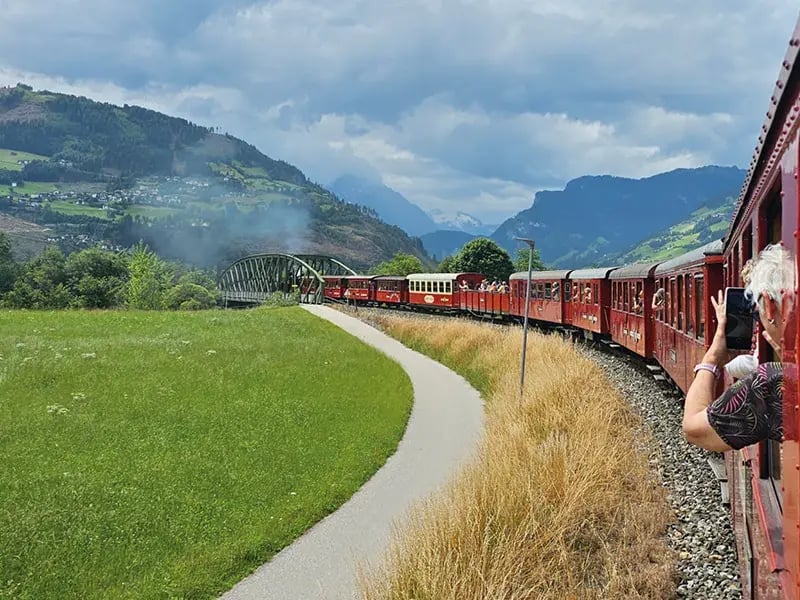 Roter Zug fährt durch grüne Landschaft mit Bergen und Brücke