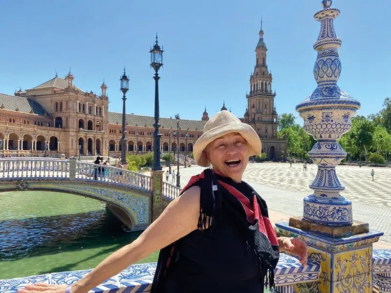 Frau mit Hut steht an verzierter Brücke vor historischer Platzanlage mit Türmen und Wasserbecken
