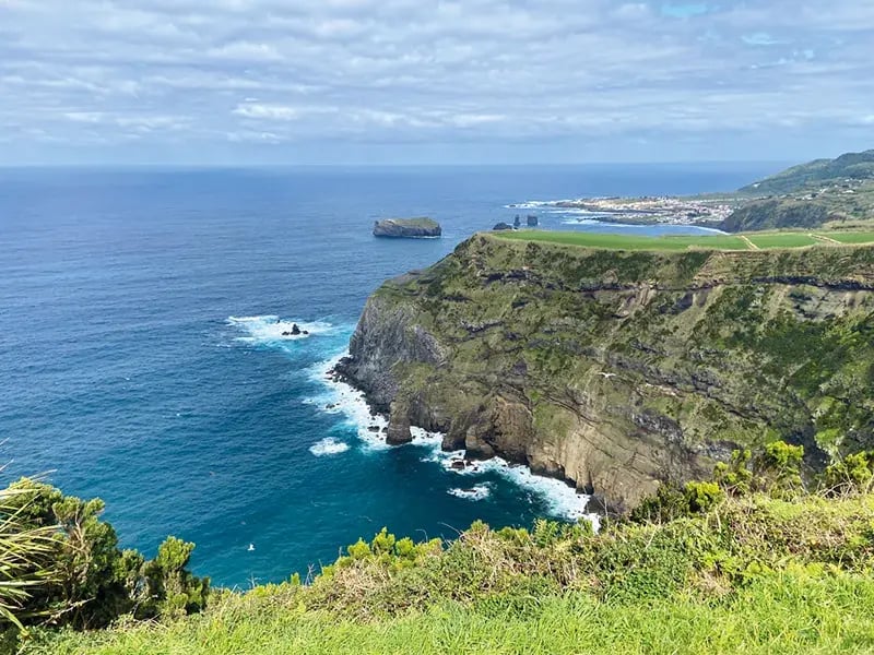 Steile Küstenklippen mit grünem Bewuchs über blauem Meer, im Hintergrund kleine Ortschaft