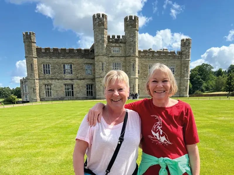 Deux femmes souriantes posent bras dessus, bras dessous sur une pelouse devant la façade en pierre du château de Leeds sous un ciel d'été.