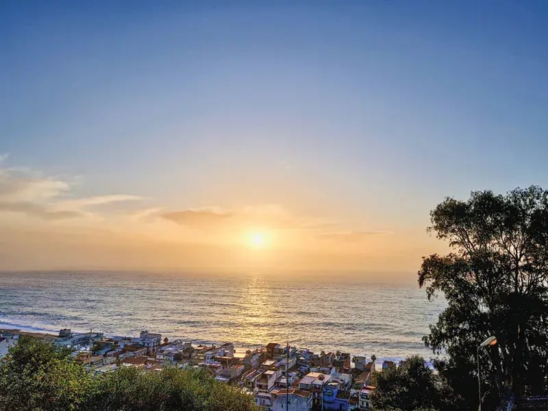 Blick von einem Hügel auf eine kleine Küstenstadt am Mittelmeer während eines goldenen Sonnenuntergangs über dem glitzernden Wasser