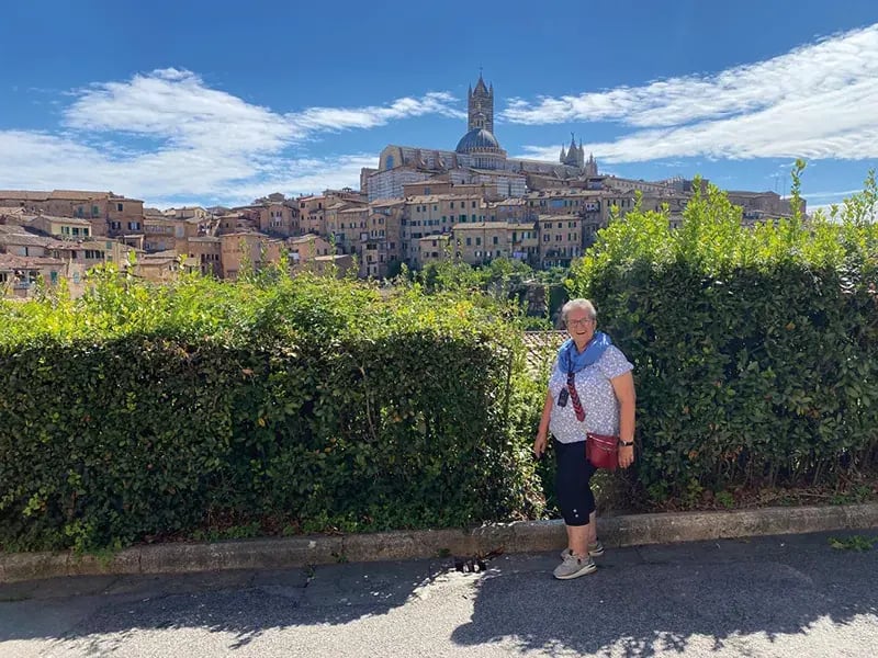 Eine lächelnde Frau steht vor einer Hecke mit dem Panorama der Stadt Siena und dem markanten Dom im Hintergrund unter blauem Himmel
