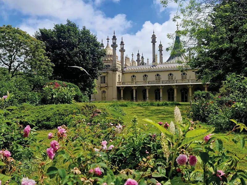 Le Royal Pavilion à Brighton vu depuis un jardin fleuri avec des roses roses sous un ciel bleu. L'architecture présente des dômes et des minarets de style indo-saracénique.