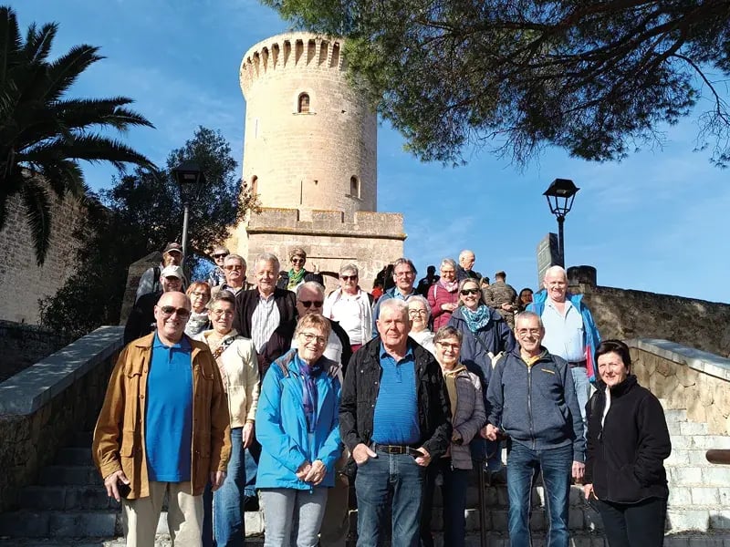 Eine große Reisegruppe posiert lächelnd auf einer Steintreppe vor einem mittelalterlichen runden Wehrturm unter strahlend blauem Himmel
