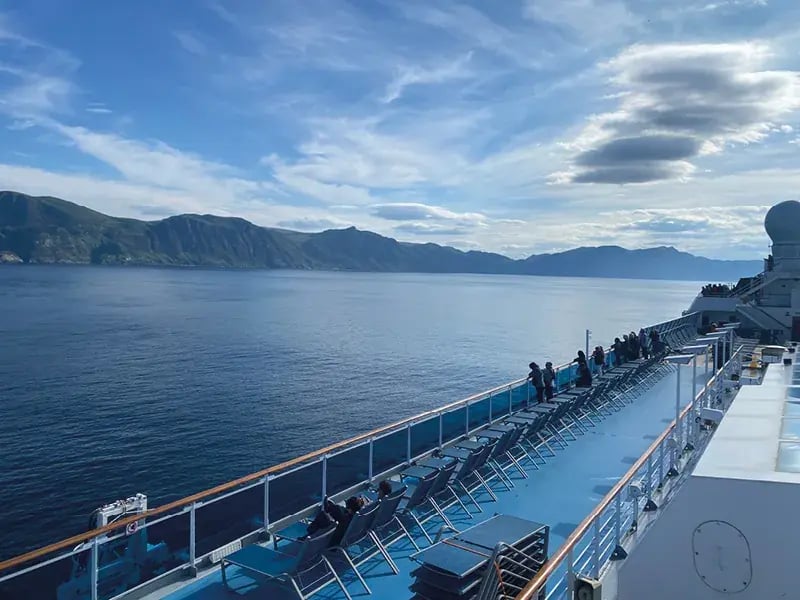 Blick vom blauen Außendeck eines Schiffes mit leeren Liegestühlen auf die weite Wasserfläche und die vorbeiziehende Gebirgsküste Norwegens