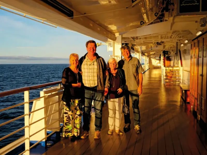 Eine Gruppe von vier Personen steht lächelnd auf dem beleuchteten Außendeck eines Kreuzfahrtschiffes bei Sonnenuntergang auf offener See.