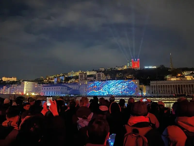 Eine Menschenmenge betrachtet bei Nacht Lichtprojektionen auf historischen Gebäuden in Lyon; die Basilika Notre-Dame de Fourvière ist rot beleuchtet
