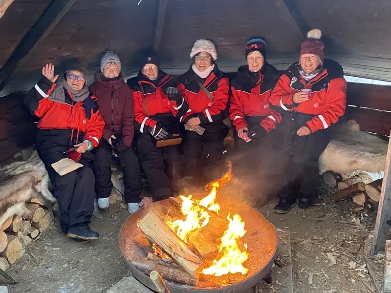 Gruppe von Personen in Winterkleidung sitzt um ein Feuer in einer Hütte