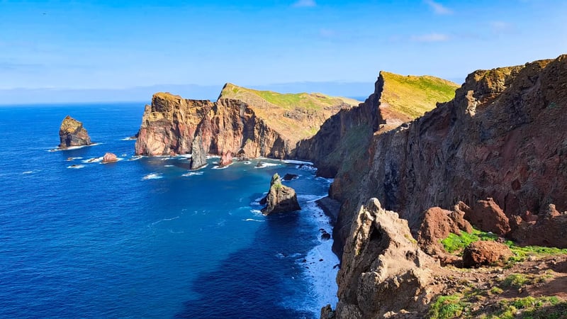 Blick von einer hohen, rötlichen Steilklippe auf den tiefblauen Atlantik mit schäumender Gischt an der felsigen Küstenlinie.