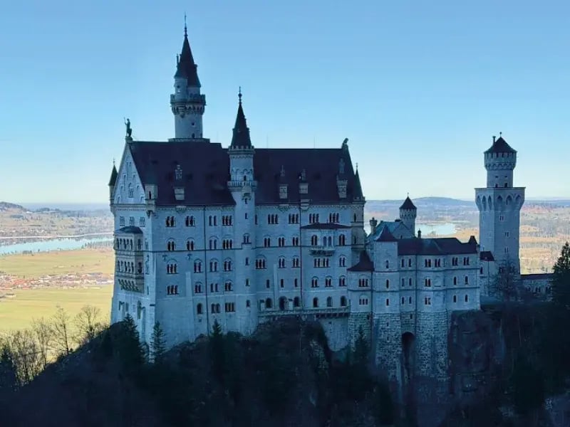 Das Schloss Neuschwanstein auf einem bewaldeten Felshügel vor einer weiten bayerischen Landschaft mit Seen unter klarem blauem Himmel