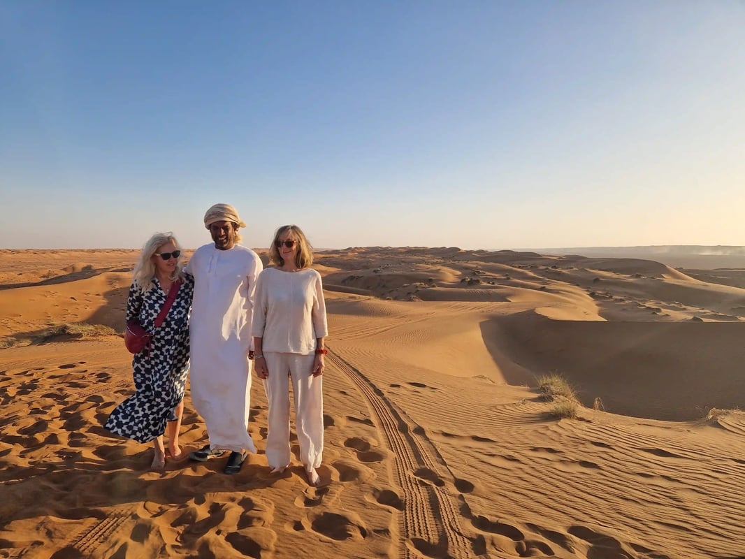 Drei Personen stehen lächelnd in einer weiten Dünenlandschaft vor einem klaren, blauen Himmel.