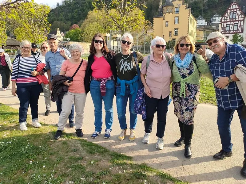 Gruppe von Menschen steht auf einem Weg im Freien, im Hintergrund Häuser und Bäume