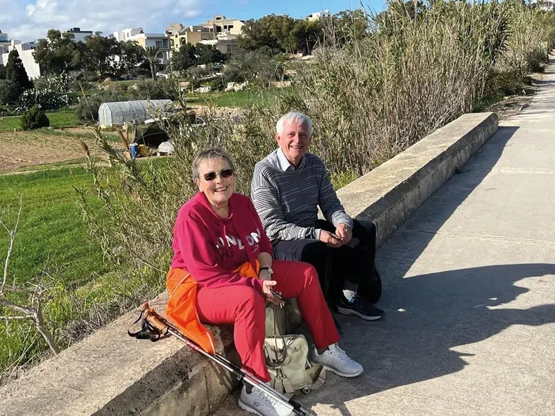 Zwei ältere Personen sitzen auf einer Mauer am Wegesrand, ländliche Umgebung mit Feldern im Hintergrund