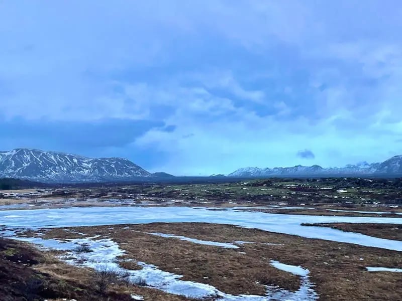 Paysage d'hiver islandais avec des étendues d'eau partiellement gelées, des prairies brunes et des montagnes enneigées sous un ciel nuageux bleuté.