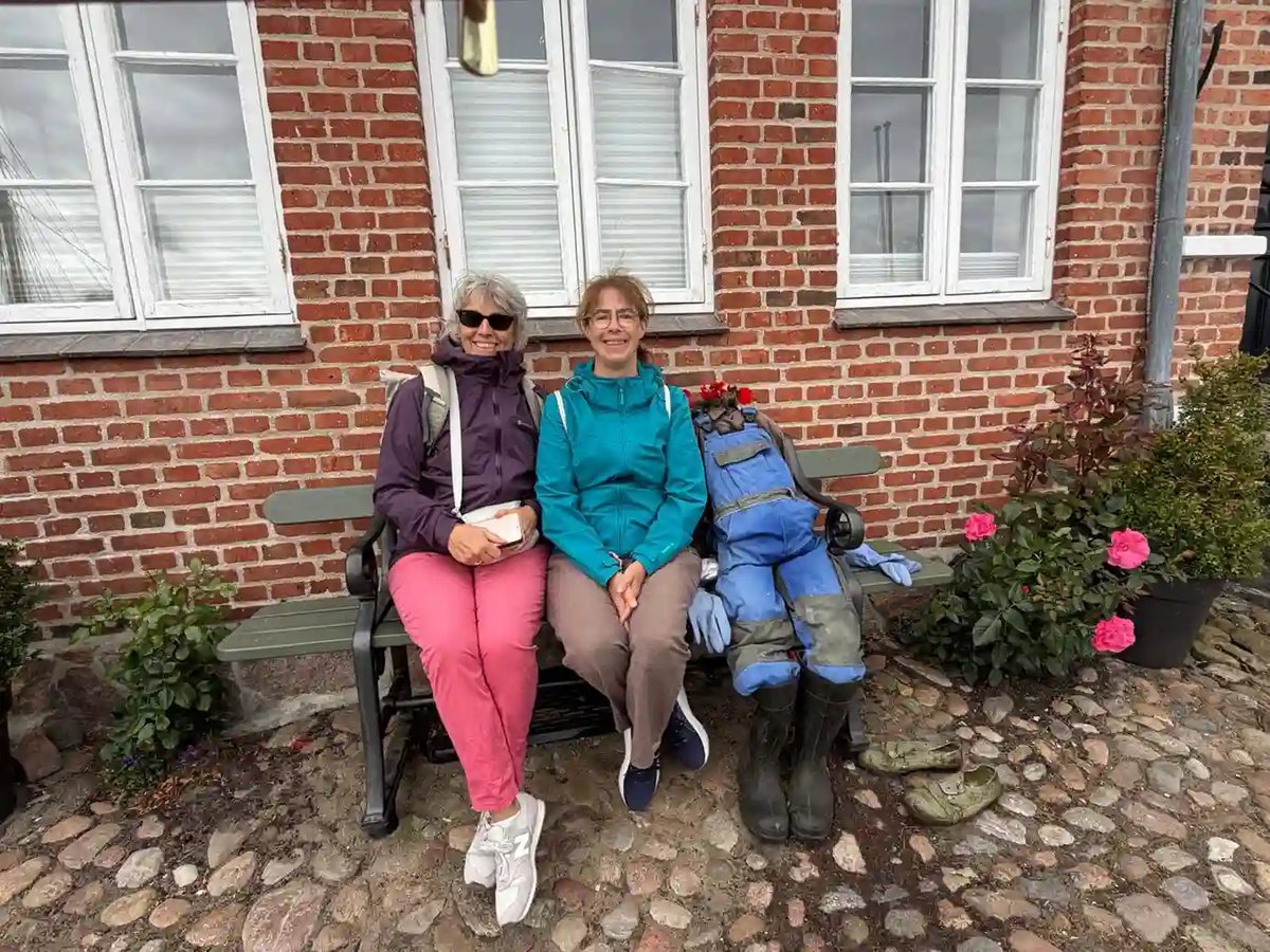 Zwei Frauen auf einer Bank neben einer dekorativen Figur vor einem Backsteinhaus