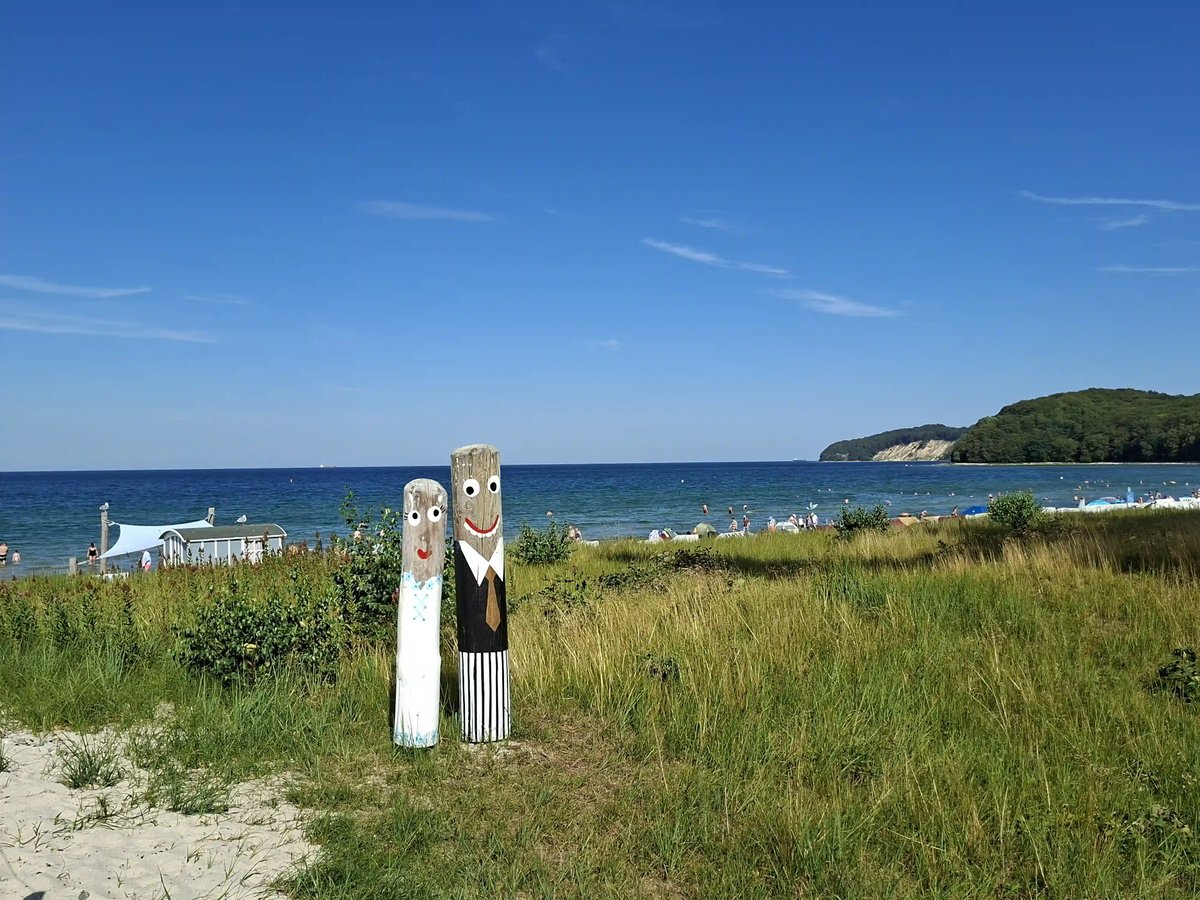 Ostseestrand mit Dünen, blauem Meer und zwei bemalten Holzfiguren im Vordergrund