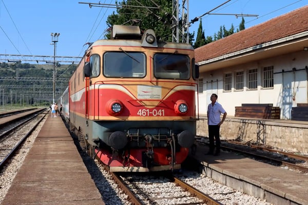 Elektrische Lokomotive steht an einem Bahnsteig in Montenegro, daneben ein Mann, Gleise und Bahnhofsgebäude im Hintergrund