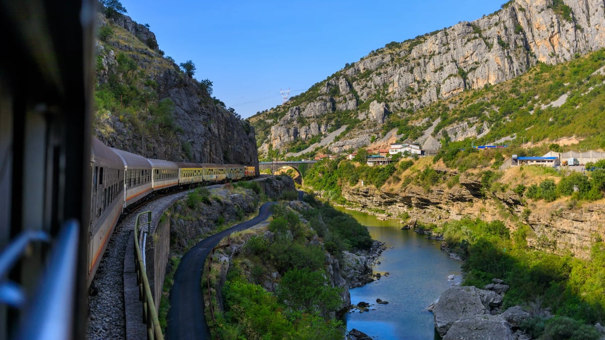 Personenzug fährt entlang einer kurvigen Strecke durch ein enges Flusstal mit steilen Felswänden und grüner Vegetation