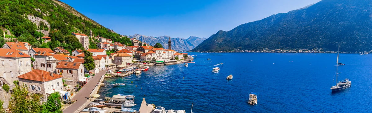 Historischer Ort mit Steinhäusern und roten Dächern direkt am Ufer der Bucht von Kotor, Boote liegen im Wasser