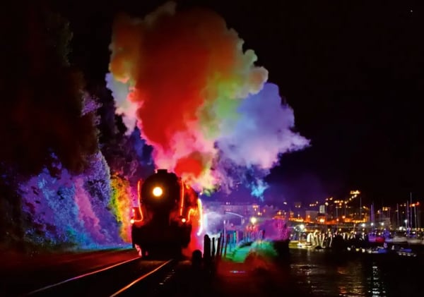 Historische Dampflokomotive bei Nacht mit farbig beleuchtetem Rauch entlang einer Bahnstrecke am Wasser