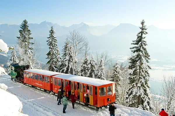 Roter Zug der Schafbergbahn steht im Schnee, während Fahrgäste in winterlicher Berglandschaft aussteigen.