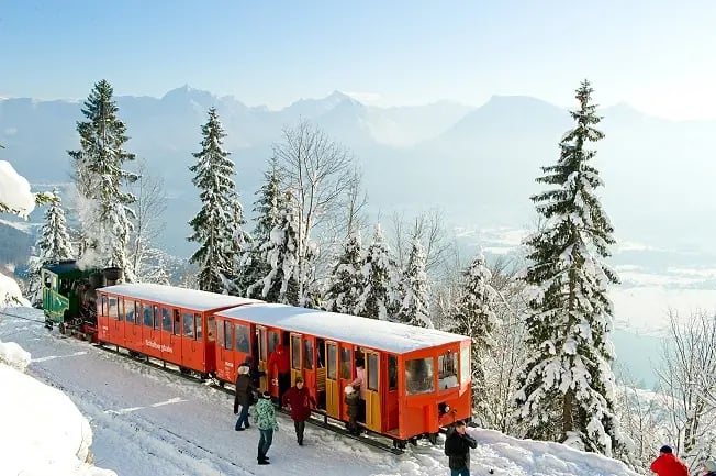 Roter Zug der Schafbergbahn steht im Schnee, während Fahrgäste in winterlicher Berglandschaft aussteigen.