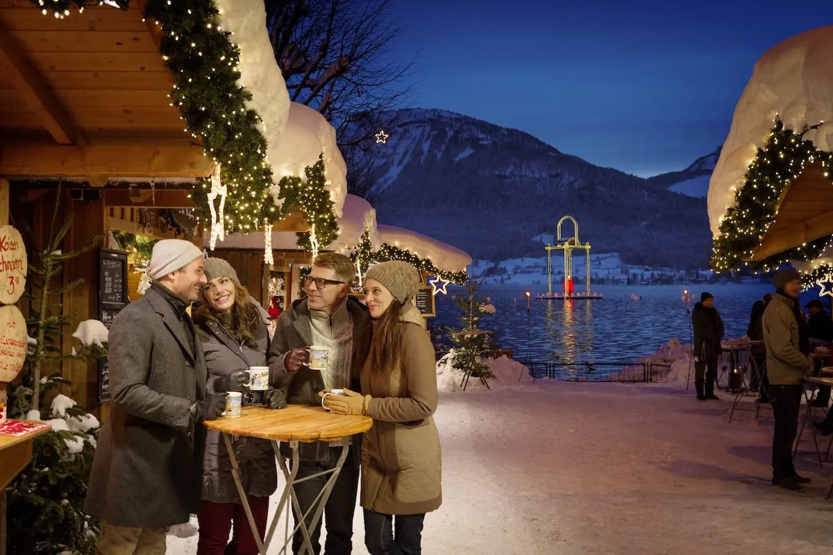 Vier Erwachsene stehen auf einem verschneiten Weihnachtsmarkt am See und trinken heisse Getränke vor beleuchteten Holzständen und winterlicher Berglandschaft.