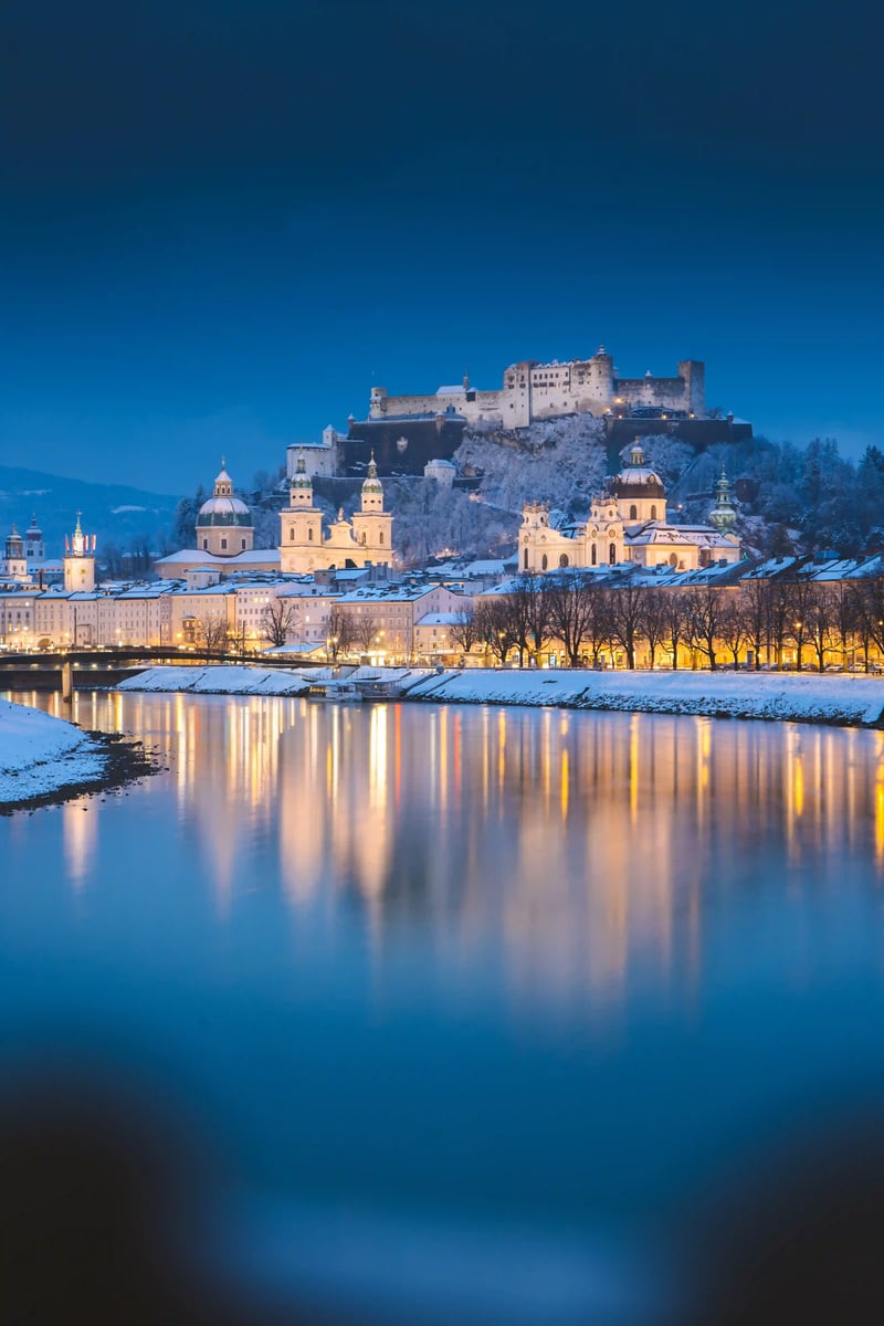 Blick auf die verschneite Altstadt von Salzburg mit beleuchteten Kirchen und der Festung auf dem Hügel bei Abenddämmerung.