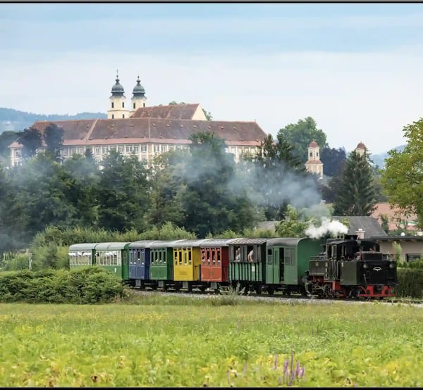 Historischer Stainzer Flascherlzug fährt durch grüne Landschaft mit Schloss im Hintergrund