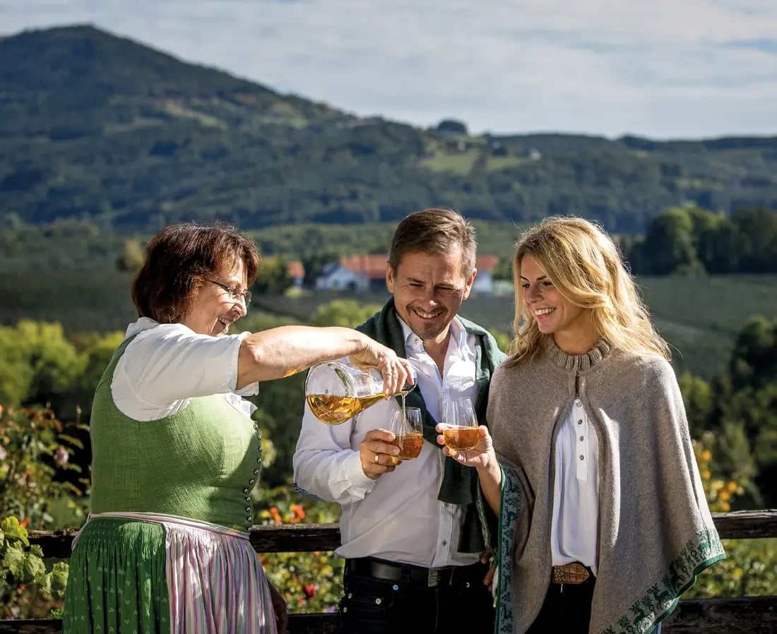 Trois personnes en tenue traditionnelle degustent des boissons devant un paysage de collines verdoyantes