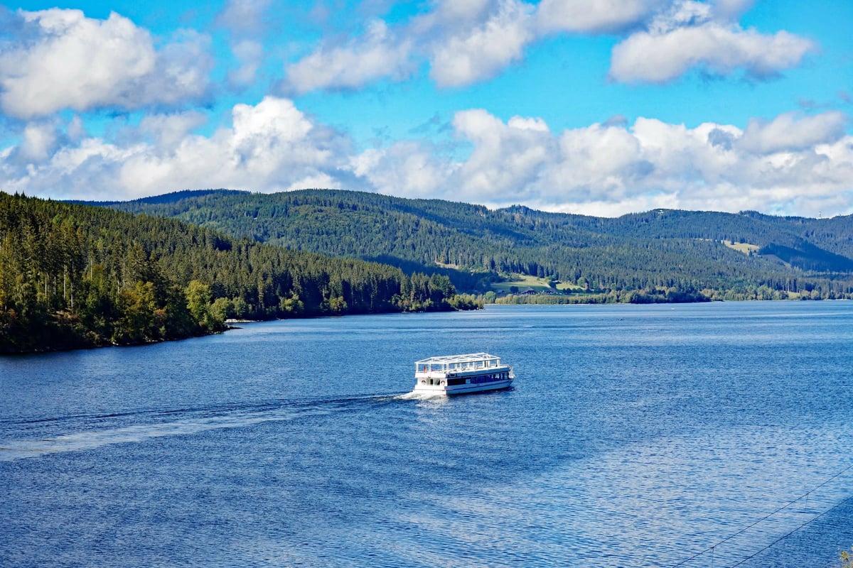 Bateau de promenade naviguant sur un lac calme entoure de forets denses et de collines sous un ciel bleu
