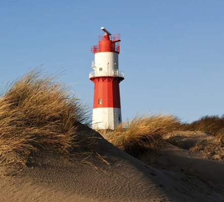 Rot weisser Leuchtturm ragt aus Sanddünen mit Strandgras vor blauem Himmel