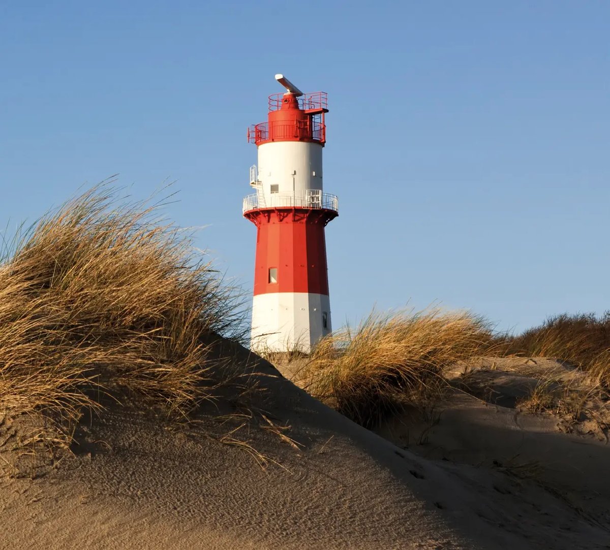 Rot weisser Leuchtturm ragt aus Sanddünen mit Strandgras vor blauem Himmel