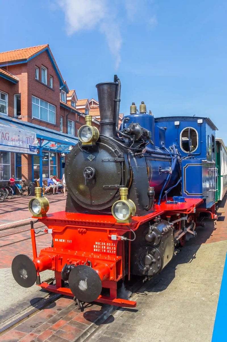 Historische schwarze Dampflok mit rotem Fahrgestell steht in einer Strasse mit Gebäuden und sitzenden Menschen