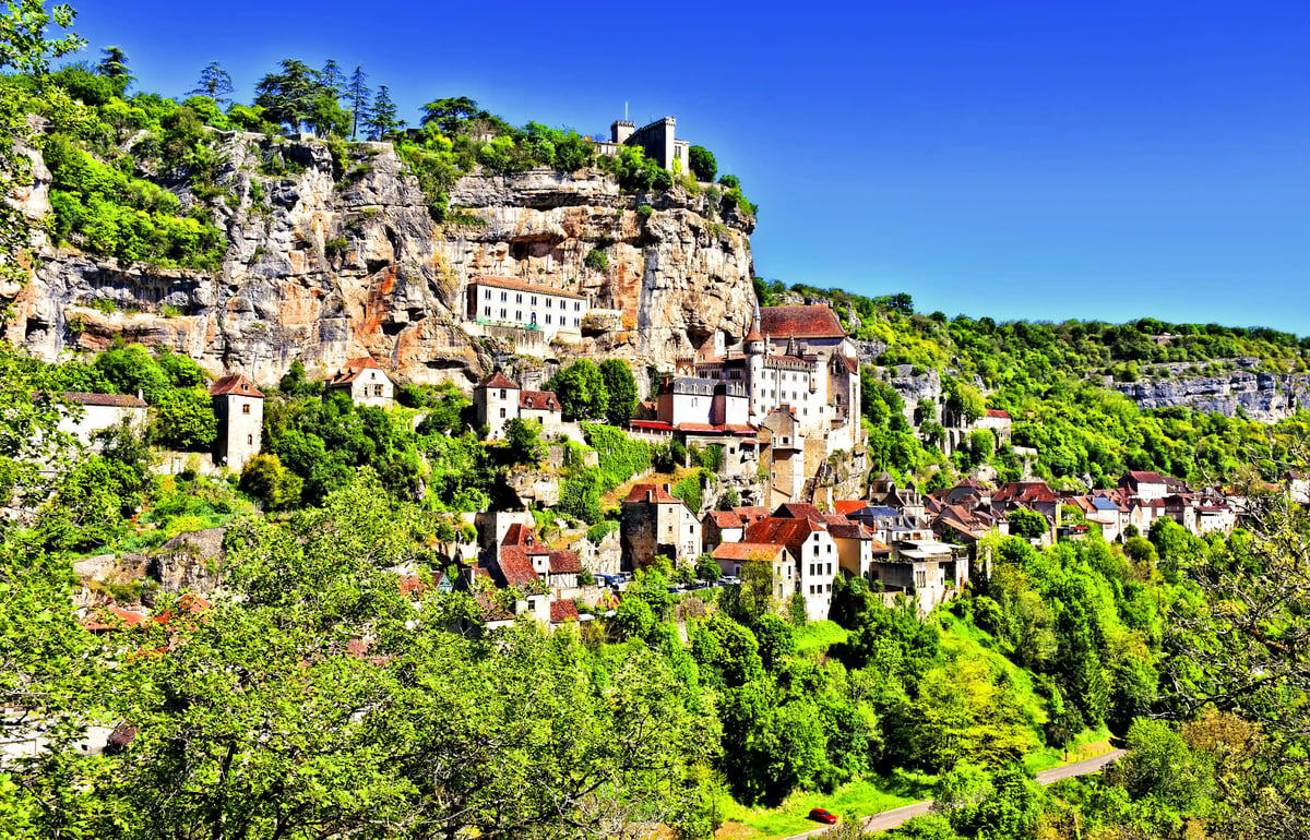 Maisons et sanctuaires médiévaux accrochés à une falaise au-dessus d'une vallée verdoyante, ciel bleu