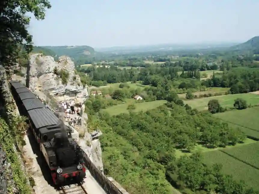 Schwarzer Zug fahrt dicht an einer Felswand entlang, darunter weites grunes Tal in der Ferne