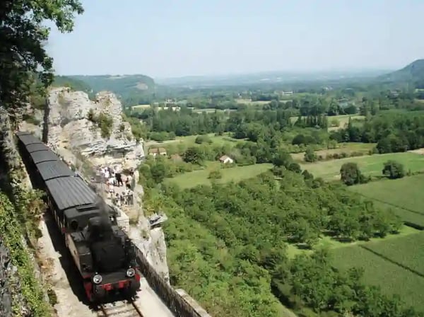 Schwarzer Zug fahrt dicht an einer Felswand entlang, darunter weites grunes Tal in der Ferne