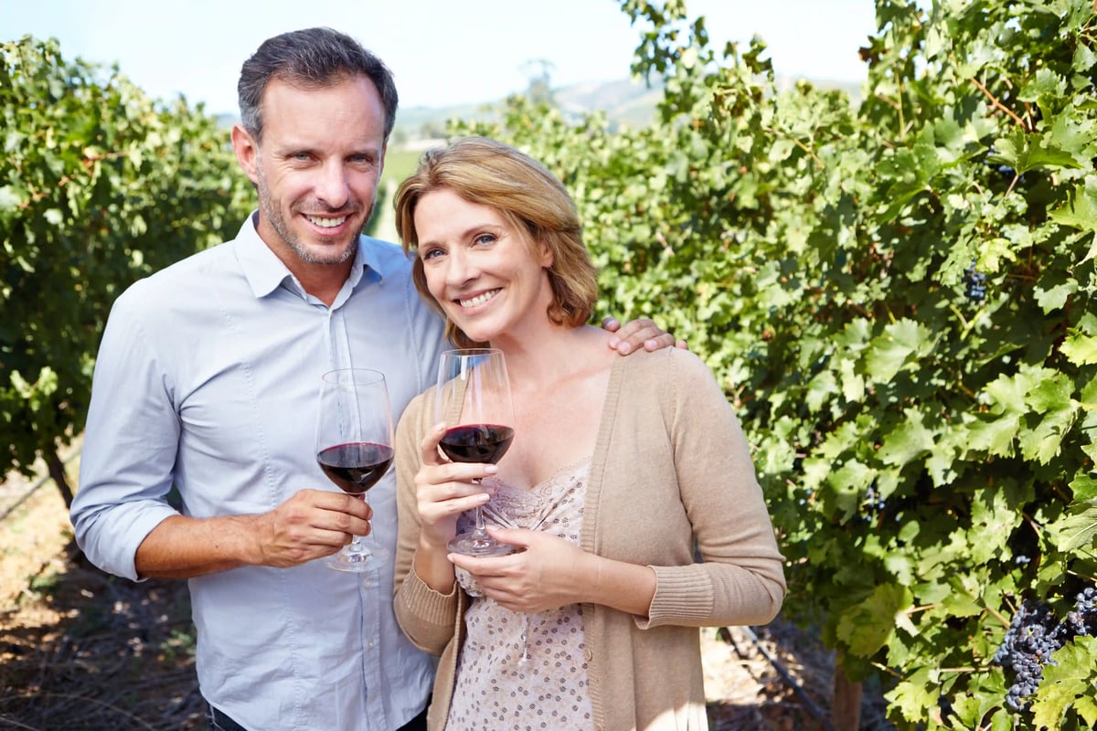 Couple dans les vignes tenant des verres de vin rouge, souriant au milieu des ceps charges de grappes