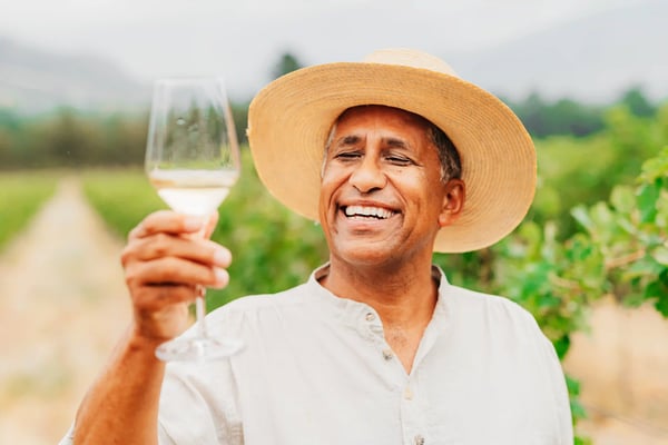 Ein Mann mit Strohhut hält lächelnd ein Glas Weisswein in einem Weinberg