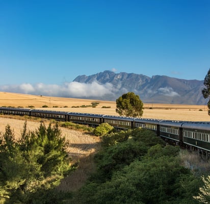 Langer, dunkler Luxuszug fährt durch weite Felder mit Bergen und tiefblauem Himmel im Hintergrund